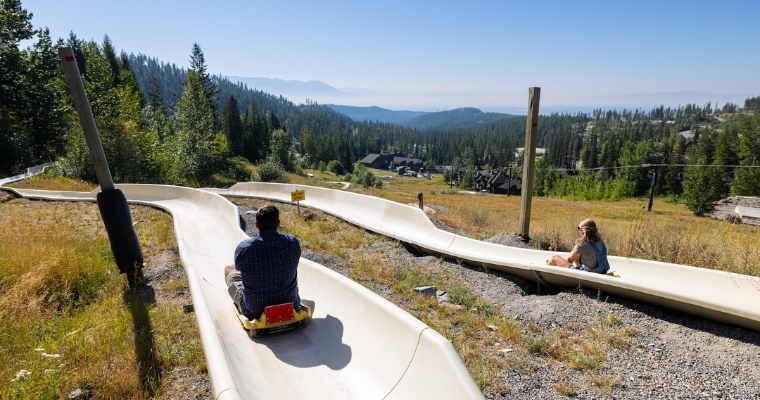 Two individuals enjoy a thrilling ride down the Alpine Slides at Whitefish Mountain Resort, surrounded by beautiful scenery.