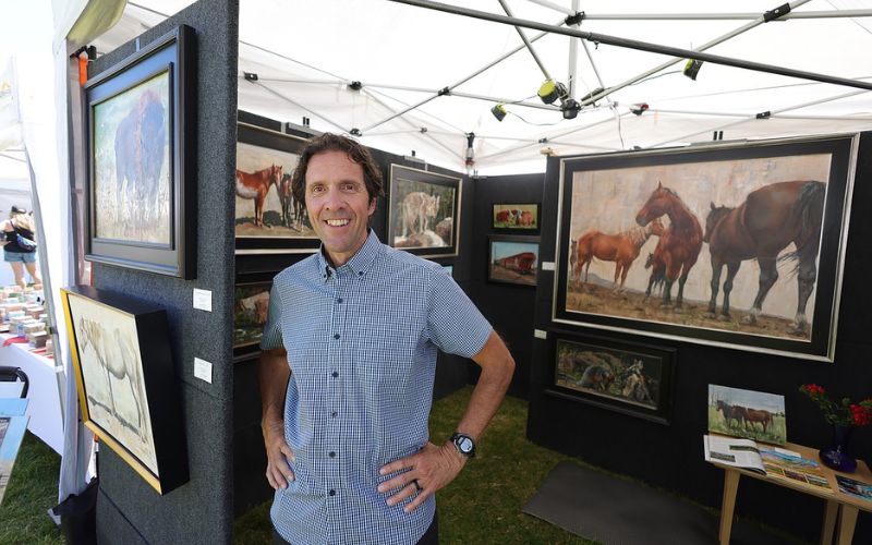 A man stands before an array of paintings at the Whitefish Arts Festival, celebrating artistic talent and creativity.