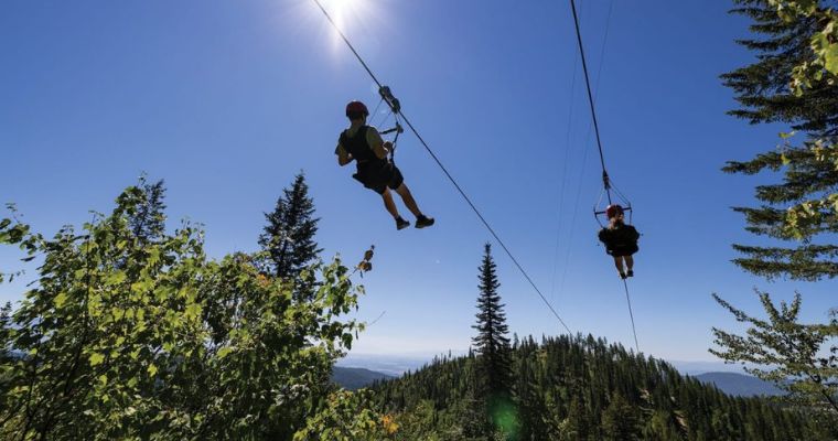 Two people experience the excitement of ziplining through the majestic mountains at Whitefish Mountain Resort, embracing adventure.