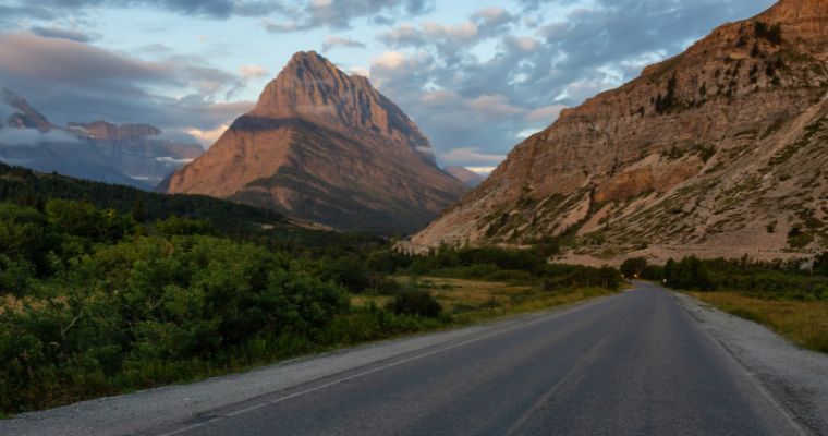 A scenic drive in Whitefish, Montana, featuring a winding road leading toward a majestic mountain peak, surrounded by cliffs and greenery at sunset.