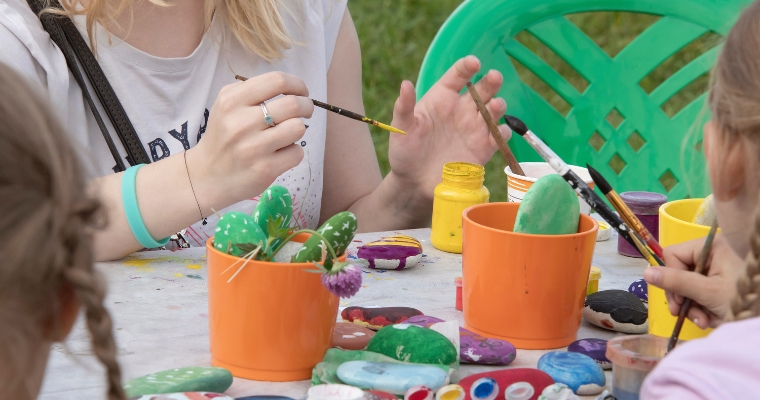 At the Whitefish Arts Festival, a woman guides children in painting a cactus at a table during an interactive art session.