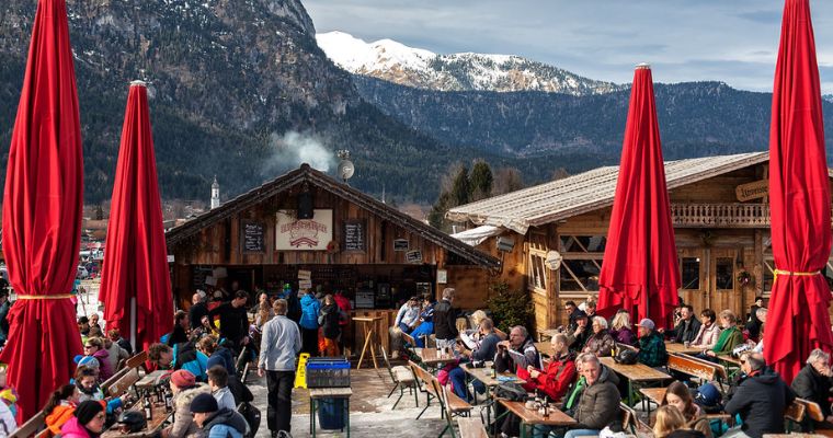 A lively après-ski scene with people socializing around a bar and restaurant in a picturesque mountain setting.