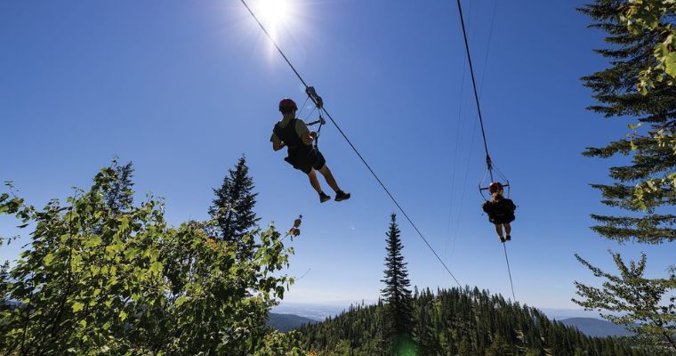 Two individuals glide on a zipline amidst the scenic mountains of Whitefish during summer.