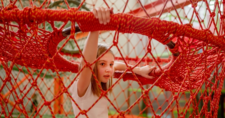 A young girl playfully sits inside a red net, surrounded by the vibrant scenery of Spider Monkey Mountain in summer.