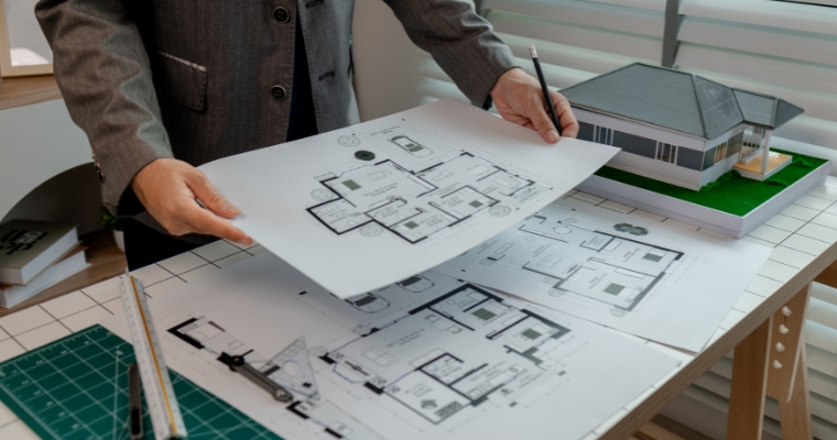 A man holds a blueprint and studies a model house, representing the construction process in Whitefish.