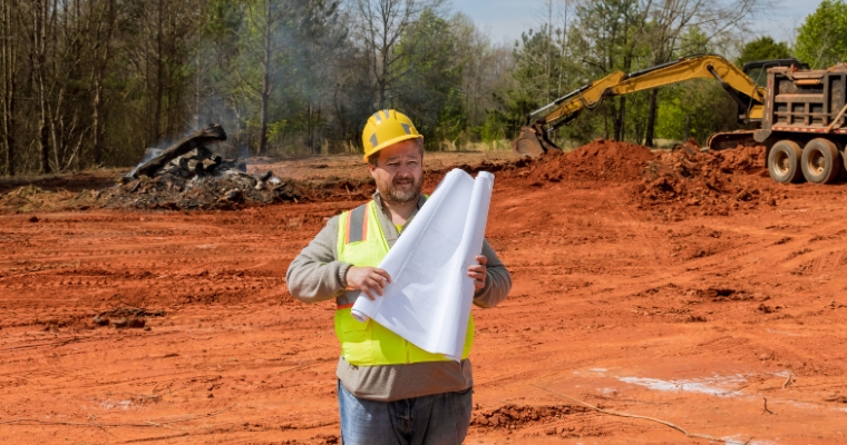 A man holds a blueprint at a construction site, focusing on site preparation for building a house in Whitefish, MT.