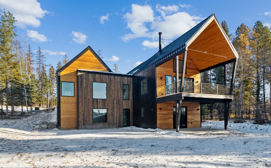 Modern cabin with wooden and dark metal siding in a snowy forest setting, illustrating the custom modern homes Whitefish.