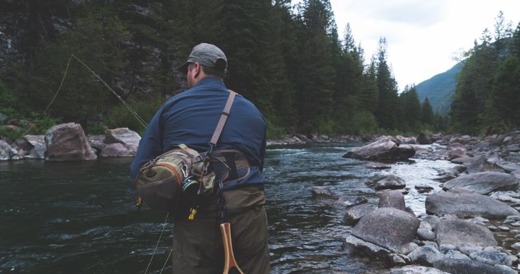 A man wades in a river, fly rod in hand, experiencing spring fishing in the beautiful landscapes of Whitefish, Montana.